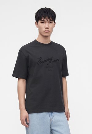 Νέος άνδρας που φοράει μαύρο T-shirt Emporio Armani και ανοιχτό μπλε τζιν, στέκεται μπροστά σε ένα απλό λευκό φόντο.