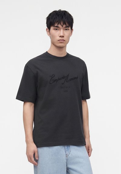 Νέος άνδρας που φοράει μαύρο T-shirt Emporio Armani και ανοιχτό μπλε τζιν, στέκεται μπροστά σε ένα απλό λευκό φόντο.