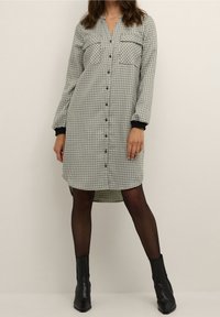 Abito camicia a quadri in bianco e nero con maniche lunghe, chiusura a bottoni e due tasche sul petto. Presenta un orlo asimmetrico e dettagli neri sui polsini.