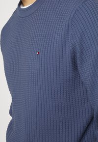 Marinefarbener strukturierter Pullover mit geripptem Muster, Rundhalsausschnitt und kleinem Logo in Rot, Weiß und Blau auf der Brust.