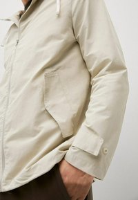 Giacca zip-up beige chiaro con colletto alzato, tasca laterale e dettaglio dei polsini con bottoni. Tessuto liscio e leggermente lucido.
