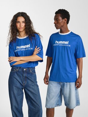 To modeller iført blå "hummel PERFORMANCE" T-shirts; den ene med lange jeans, den anden med lyse denimshorts, stående mod en hvid baggrund.