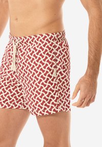 Costumi da bagno rossi e crema con motivo, dotati di vita elastica e cordoncino. Presentano un design ondulato su tutta la superficie del tessuto. Nessuna tasca visibile.