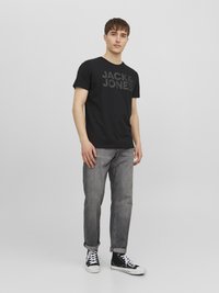 Black cotton t-shirt with "JACK & JONES" logo in dark gray, paired with gray jeans and black high-top sneakers. Casual fit and simple design.
