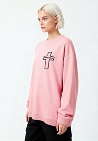 Femme aux cheveux blonds courts portant un sweat-shirt rose oversize avec une croix noire et le texte "KARMA KILLS", associée à un pantalon noir, regardant sur le côté.