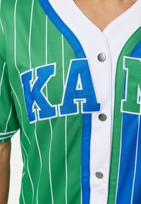 Žalias ir mėlynas dryžuotas beisbolo džersis su baltomis detalėmis, ant kurio didelėmis baltomis raidėmis užrašyta „KAM“, o priekyje yra spaudiniai su mygtukais.