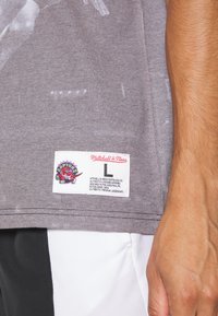 Grå bomulds t-shirt med et stort grafisk print, et mærke med "Mitchell & Ness" og et Raptors-logo patch nær kanten.