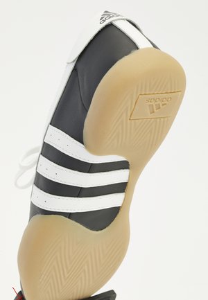 Sort Adidas sneaker med hvide striber og gummisål, der viser Adidas-logo på sålen, vinklet opad mod hvid baggrund.