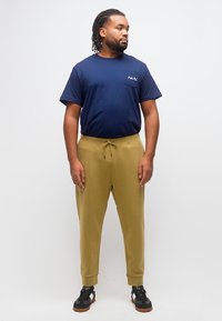 Άνδρας με πλεγμένα μαλλιά και γενειάδα, φοράει μπλε ναυτικό T-shirt και χακί φόρμες, στέκεται μπροστά σε απλό ανοιχτό φόντο.