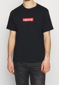 Mężczyzna w czarnej koszulce z krótkim rękawem z czerwonym logo Levi's umieszczonym na środku klatki piersiowej, w połączeniu z ciemnoszarymi dżinsami.