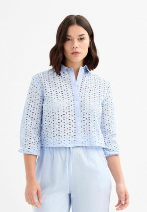 Camisa de botón recortada de color azul claro, con diseño de ojetes, mangas de tres cuartos y una textura suave. Presenta un cuello clásico y botones en la parte delantera.