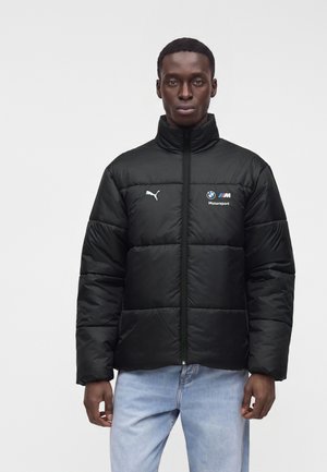 Homme portant une doudoune noire Puma BMW Motorsport et un jean bleu clair sur un fond blanc uni.