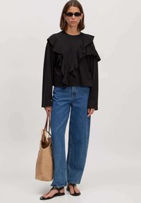 Zwarte blouse met lange mouwen en gerimpelde details, gecombineerd met hooggetailleerde blauwe spijkerbroeken. Accessoires: geweven tote tas en zwarte sandalen.