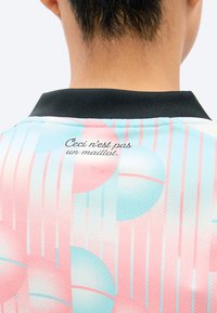 Persoană purtând o cămașă pastelată roz și albastră cu model și guler negru, text în franceză pe partea superioară a spatelui care spune „Ceci n'est pas un maillot”.