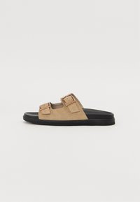 Beige semskede sandaler med to justerbare stropper og metallspenner på en svart gummisåle. Flat design, myk tekstur, åpen tå-stil.