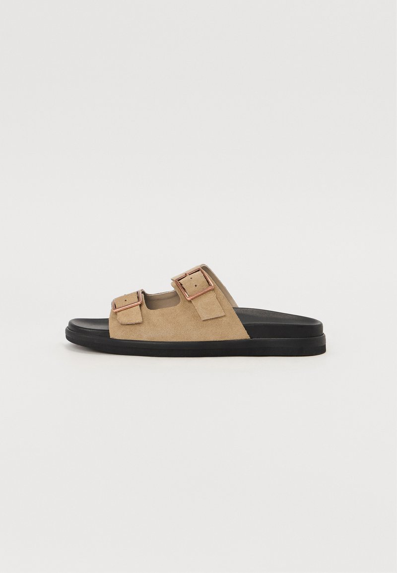 Beige semskede sandaler med to justerbare stropper og metallspenner på en svart gummisåle. Flat design, myk tekstur, åpen tå-stil.