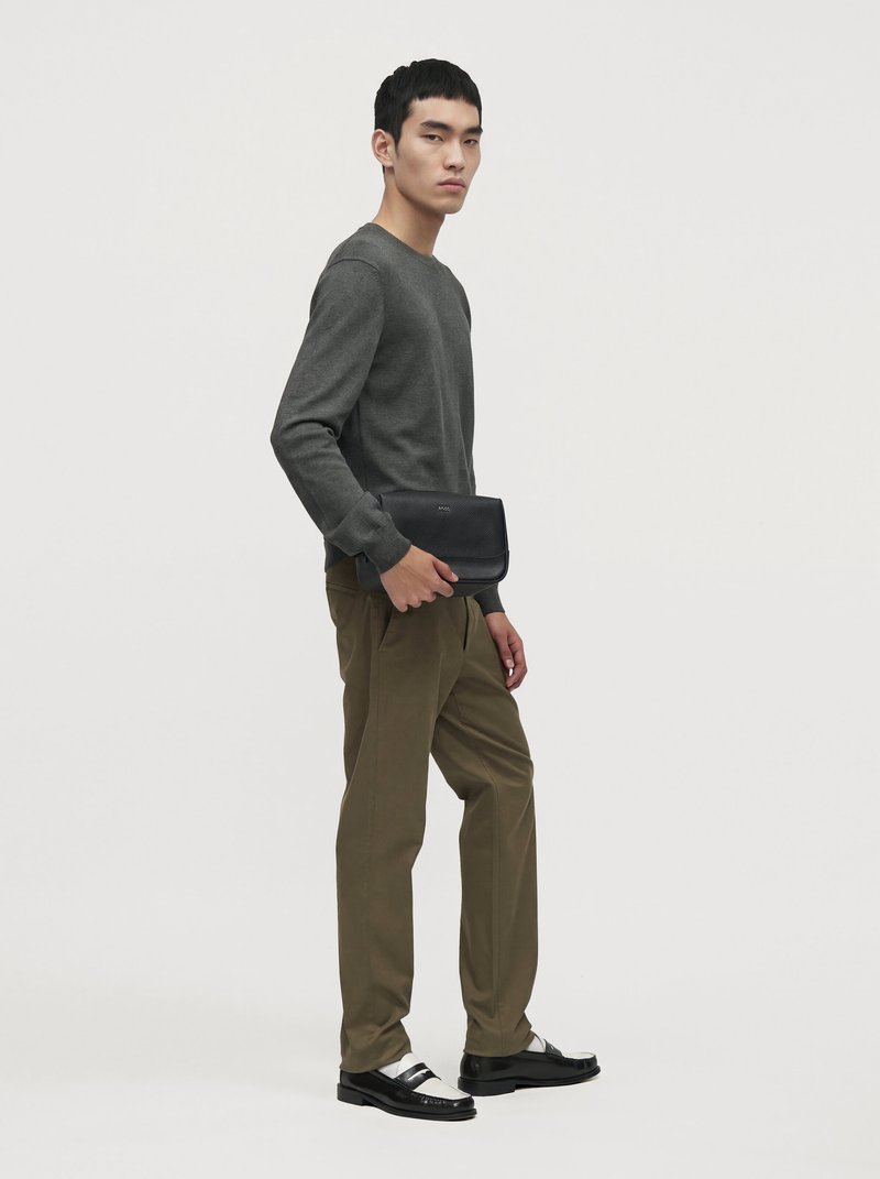 Un homme en pull gris anthracite et pantalon olive tient une pochette noire et se tient devant un fond clair uni.