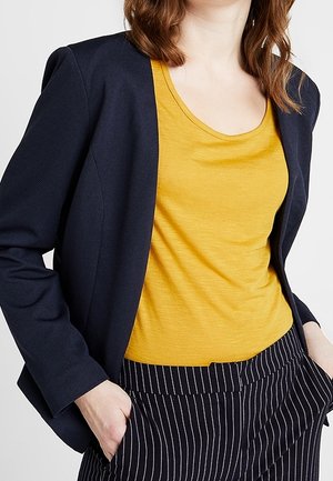 Vrouw met een mosterdgele top met ronde hals, een marineblauwe blazer en marineblauwe gestreepte broek, handen in de zakken, torso zichtbaar.