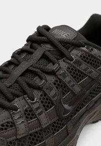 Musta verkkokenkä tummanharmailla kumiyksityiskohdilla, paksuilla nauhoilla ja näkyvällä Nike-logolla. Sisältää teksturoituja päällysteitä ja tukevan kehikkosuunnittelun.
