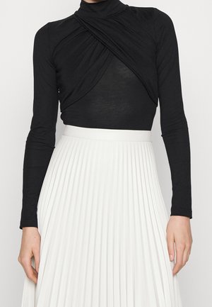 Femme portant un haut noir à manches longues avec un détail torsadé à l’avant et une jupe blanche plissée taille haute.