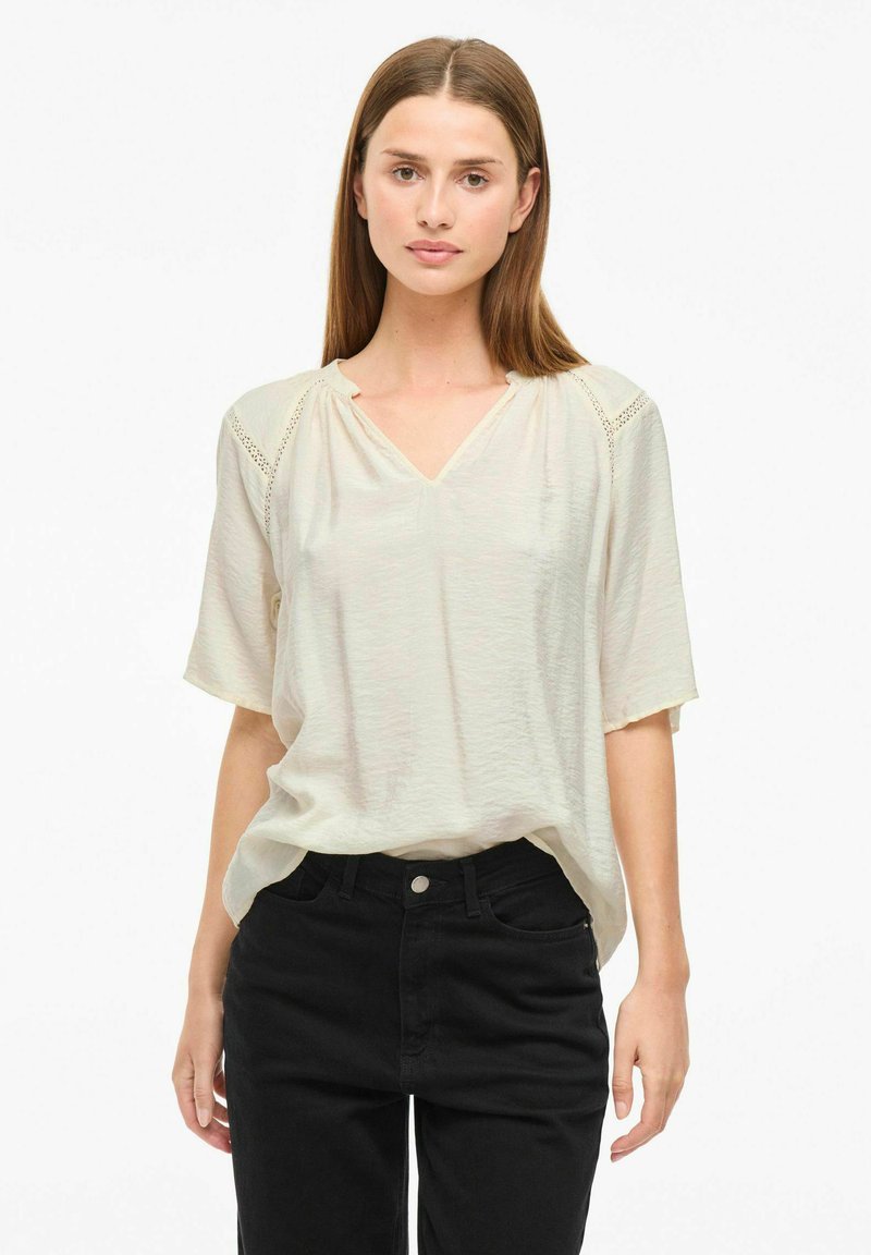 Gräddfärgad kortärmad topp med V-hals och spetsdetaljer på axlarna, ihop med svarta högmidjade jeans. Mjuk, lättviktsmaterial.