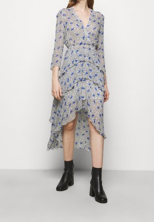 Vrouw draagt een witte en blauwe bloemenwikkeljurk met een gerimpelde asymmetrische zoom, gecombineerd met zwarte enkellaarsjes, tegen een effen achtergrond.