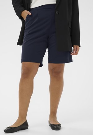 Person iført marineblå knælange shorts, sort blazer, hvid top og sorte flade sko, stående med den ene hånd i lommen på hvid baggrund.