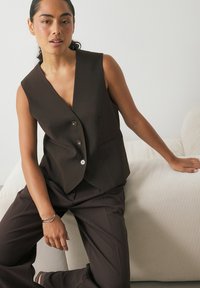 Rjava brezrokavna jopica z globokim V-izrezom, opremljena z gumbom za zapiranje in dvema sprednjima žepoma, v kompletu z usklajenimi krojenimi hlačami.