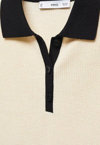 Polo shirt crème avec un col côtelé noir, équipé de trois boutons noirs et d'un tissu tricoté texturé. Étiquette de la marque visible à l'intérieur du col.