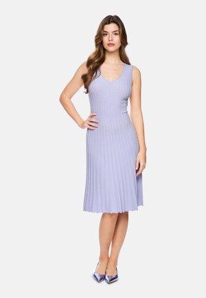 Frau mit langen braunen Haaren, die ein ärmelloses, knielanges lavendelfarbenes plissiertes Kleid und metallisch violette spitze Schuhe trägt, steht mit einer Hand in der Hüfte.