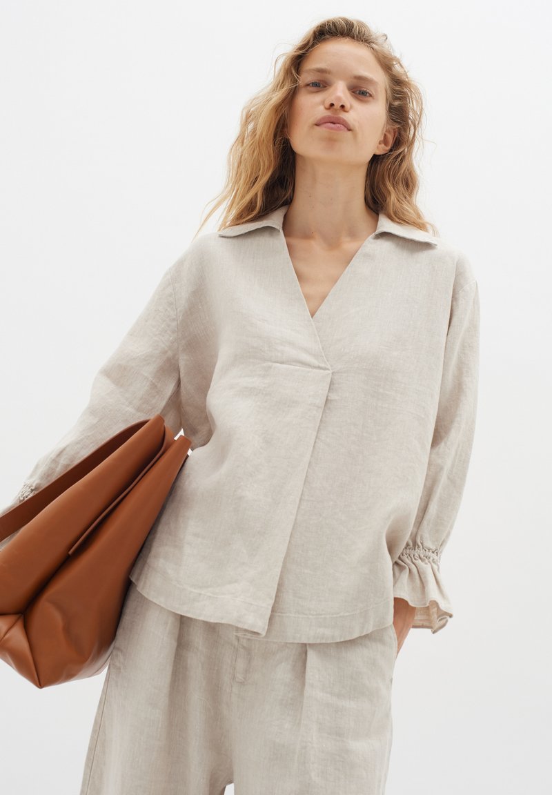 Blusa in lino beige con maniche lunghe, scollo a V e polsini arricciati. Indossata con pantaloni chiari e una borsa in pelle marrone.
