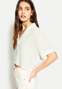 Blusa bianca corta realizzata in tessuto leggero con un motivo testurizzato sottile, dotata di un'apertura frontale con bottoni e maniche corte arrotolate.