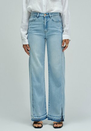 Mujer que lleva vaqueros de tiro alto y pierna ancha en azul claro con aberturas en el dobladillo, combinados con una camisa blanca abotonada y sandalias negras de punta abierta.
