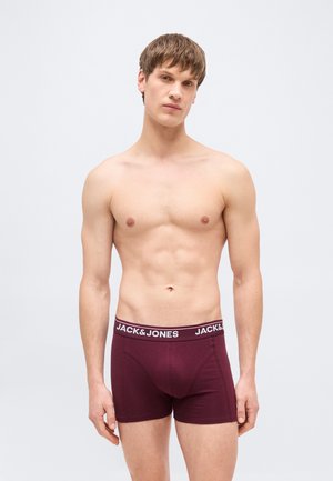 Jeune homme debout, torse nu, portant un boxer bordeaux avec une ceinture "JACK&JONES", sur un fond gris clair uni.