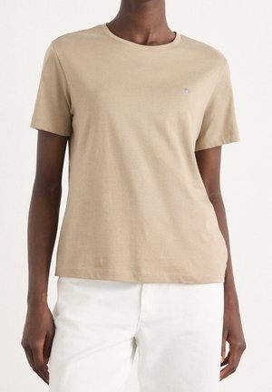 Persona que lleva una camiseta beige de manga corta con cuello redondo y pantalones blancos, de pie contra un fondo claro y liso.