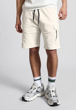 Férfi törtfehér cargo rövidnadrágot visel fekete húzózsinórokkal, fehér, zöld csíkkal díszített zoknit és ezüst színű sportcipőt.