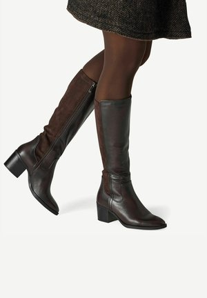 Femme portant des bottes hautes en cuir et suède marron foncé avec talons blocs et collants foncés, marchant avec une jambe levée.