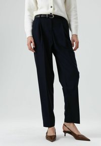 Marineblauwe hoge taillebroek met getailleerde plooien, gemaakt van gladde stof, gecombineerd met bruine puntige slingback-hakken en een witte cardigan.