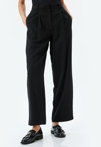Pantalones anchos y negros de tejido suave, con pliegues frontales y bolsillos laterales. Combinados con mocasines negros brillantes que presentan flecos y un detalle dorado.