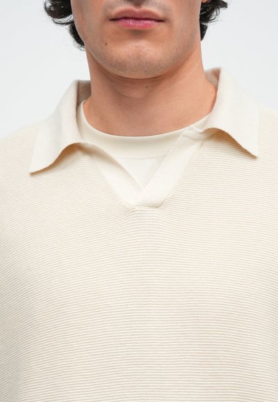 Bež pleteni pulover z V-izrezi in ravnim ovratnikom. Tkanina ima teksturiran rebrast vzorec, nanesen čez svetlo barvno srajco.