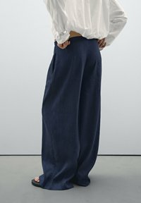 Pantalons larges en marine en tissu doux, avec des poches latérales, une coupe décontractée et une texture lisse, portés avec des sandales noires.