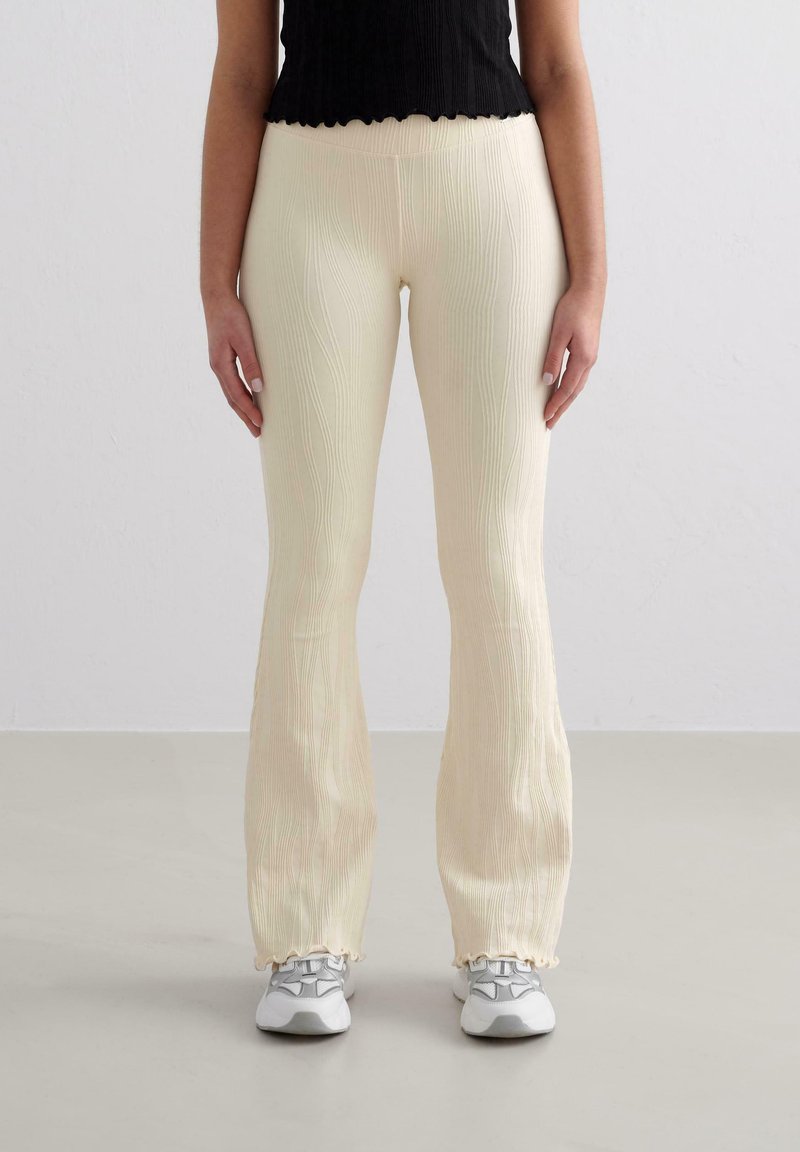 Pantalons crème côtelés et évasés avec une texture ondulée et un ourlet volanté, associés à des baskets grises et un haut noir.