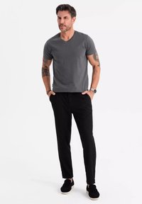 Šedé tričko s výstřihem do V, černé zúžené kalhoty a černé boty na slip. Model má tetování a stojí s rukama v kapsách na bílém pozadí.