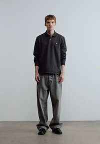 Μακρυμάνικο πουκάμισο πόλο σε σκούρο γκρι, τζιν παντελόνι σε ανοιχτό γκρι χρώμα με χαλαρή εφαρμογή και μαύρα παπούτσια φορεμένα από έναν άνδρα που στέκεται μπροστά σε απλό φόντο.