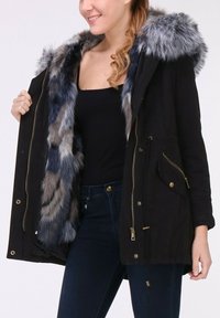 Parka noire avec une capuche doublée en fausse fourrure gris-bleu, fermeture éclair à l'avant, poches latérales et taille à cordon. Tissu lisse avec une finition légèrement texturée.