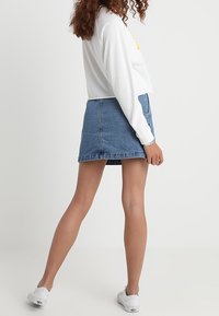 Džínová sukně s těsným pasem a rozšířeným lemem, zkombinovaná s bílým topem s dlouhým rukávem. Bílé tenisky dokončují volný vzhled.