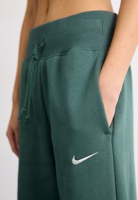 Grønne joggebukser laget av mykt stoff, med en bred ribbet midje, side lommer og en hvit Nike-logo på venstre underben.
