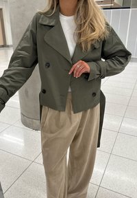 Olivgrüne doppelt-knopfbare Jacke mit großen schwarzen Knöpfen, kombiniert mit cremefarbenen, weit geschnittenen Hosen. Glatter, wasserabweisender Stoff.