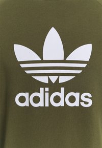 Sudadera verde oliva con un gran logo blanco de Adidas con tres rayas y un gráfico floral centrado en la parte trasera.