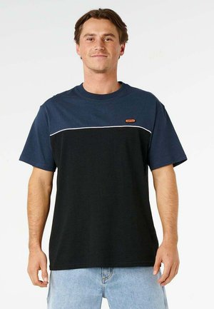 Mann trägt ein dunkelblau-schwarzes T-Shirt mit kleinem Logo und hellblauer Jeans, steht vor weißem Hintergrund.